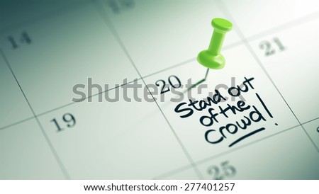 Concept image of a Calendar with a green push pin. Closeup shot of a thumbtack attached. The words Stand out of the crowd written on a white notebook to remind you an important appointment.