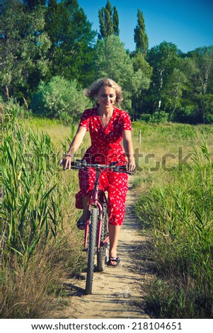 The photo shows a woman in a red sundress, she rides a bicycle in the countryside.