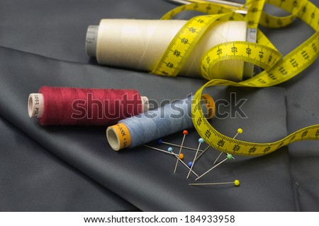 Tailors tools - thread spools, pin and yellow measuring tape on grey fabric.