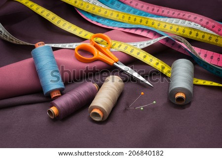 Tailors tools - thread spools, pin, scissors and measuring tape on red fabric.