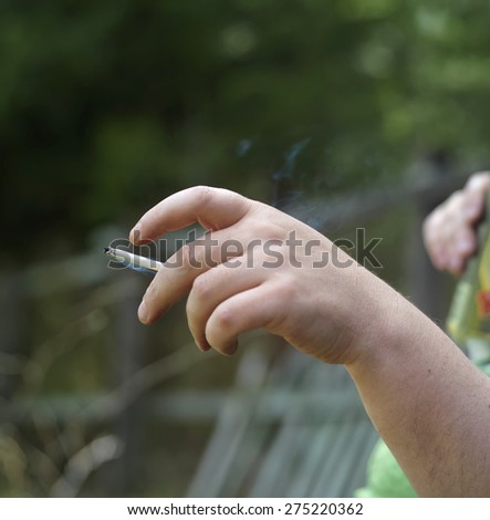 One hand with a cigarette, another holding can with drinks in the blurred background, outdoor square shot