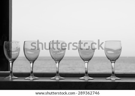 Empty wine glasses on a window sill, sea and sky in the blurred background, in black and white