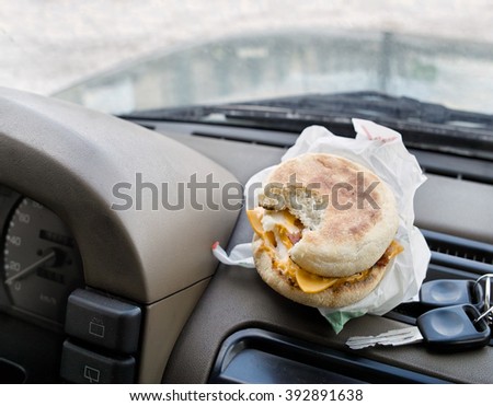 A bitten sandwich next to the ignition keys laid on a car dashboard, concept of having a coffee break while travel by car