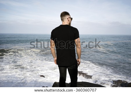 young hipster man wearing a black blank t-shirt on the ocean background. Back view