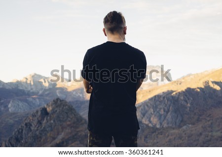young hipster man wearing a black blank t-shirt on the mountain background. Back view