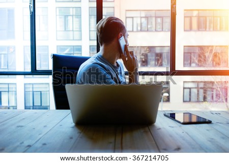Young businessman busy working on laptop computer while talking on the smart-phone at office.young hipster using laptop sitting at wooden table of modern loft space and looking out the window.flare