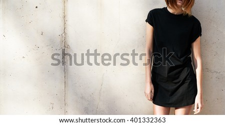 Cropped image on a stylish young woman dressed in a black blank t-shirt standing against a background of a concrete wall in the rays of the setting sun