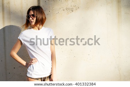 Lovely young girl wearing in a white blank t-shirt and sunglasses posing against a background of a concrete wall in the rays of the setting sun