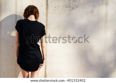 Attractive young girl wearing in a black blank t-shirt  posing against a background of a concrete wall in the rays of the setting sun.back view