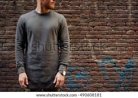 A cropped photo of a bearded hipster guy wearing blank gray long sleeve t-shirt while standing on a brick wall background on a street. Empty place for you logo or design.