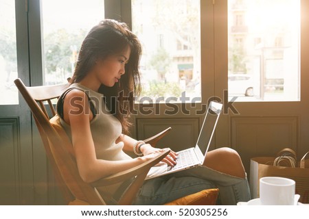Attractive asian female is typing messages on a laptop while sitting against the wide windows of a coffee shop. a hipster girl is working on a portable computer in a cafe.