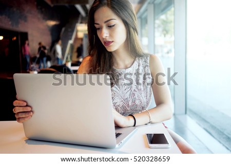 Young attractive woman is sitting in front of an open laptop in a coffee shop. Modern young business woman is working on a new project, looking at the screen of a portable computer.