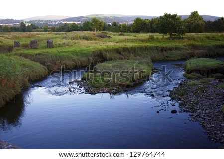 Bend in a canal in Neath with small grassy island in middle of canal.  Stones and shingle on the bank and mountains in the background