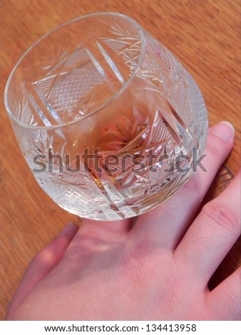Crystall glass filed with a sip of liquor and hold by the female hand. Wooden background./Crystal glass