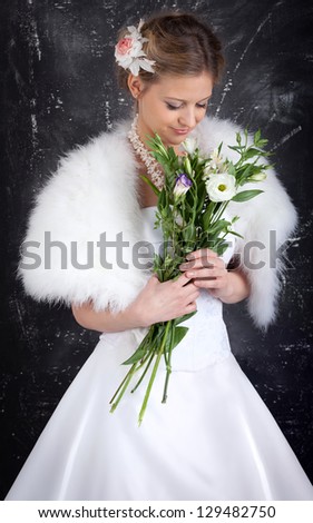 The young beautiful woman in white wedding dress and white fur mantlet watch at the bouquet of flowers in her hands. Hairdress is decorated with rose