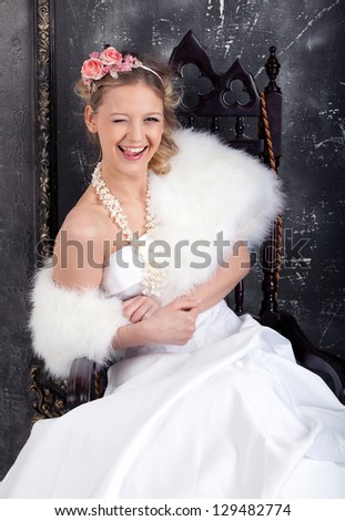 The young playful woman in white wedding dress and white fur mantlet sits on the chair.  Hairdress is decorated with pink flower composition