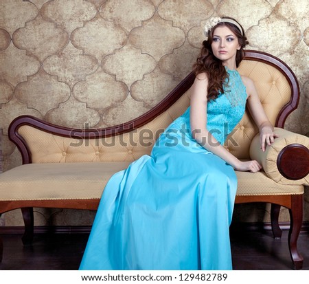 The young woman in blue evening dress sits on the sofa. Brown loose hair decorated with  beige flowers