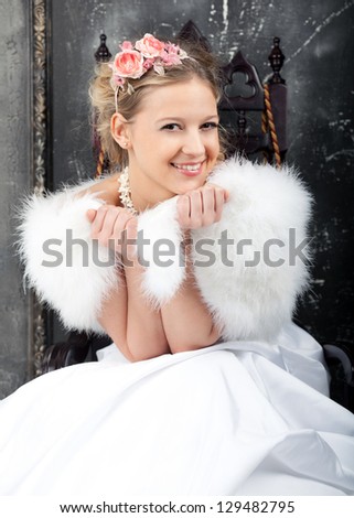 The young beautiful woman in white wedding dress and white fur mantlet sits on the chair.  Hairdress is decorated with pink flower composition