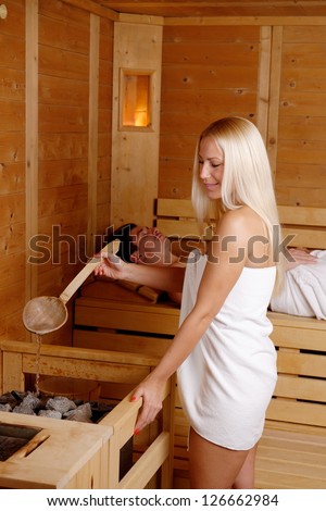 Beautiful blond girl enjoying in the sauna and puring water on the hot stones