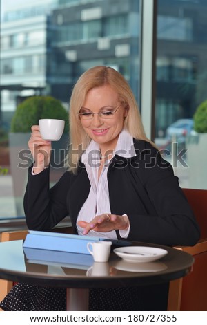 Candid photo of a attractive middle-aged blond businesswoman working at hotel lobby with a tablet pc/smartphone and drinking coffee