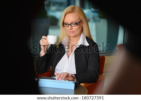 Candid photo of a attractive middle-aged blond businesswoman working at hotel lobby with a tablet pc/smart-phone and drinking coffe