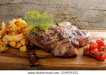 Grilled T-bone steak seasoned with spices and fresh herbs served on a wooden board with fresh tomato and roast potatoes