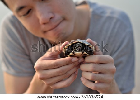 Thai young man holding a thai snail-eating turtle in his both hands with a curious face.