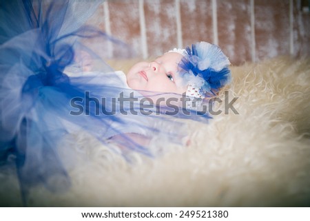 baby lying on the carpet, the little girl in a blue skirt and a blue hat on a white fluffy carpet in the room. The child in the interior