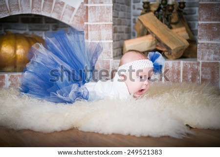 baby lying on the carpet, the little girl in a blue skirt and a blue hat on a white fluffy carpet in the room. The child in the interior