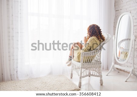 girl drinking coffee or tea in morning sunlight near window. girl in a knitted jacket looking through a window, sitting back, drinking tea and sad, white interior of the house