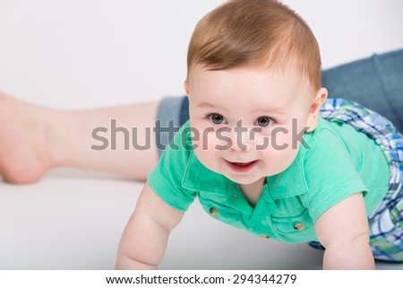 8 month year old baby crawls towards camera, while mom leg is sitting in background. dressed in a cute green polo shirt and blue plaid shorts.