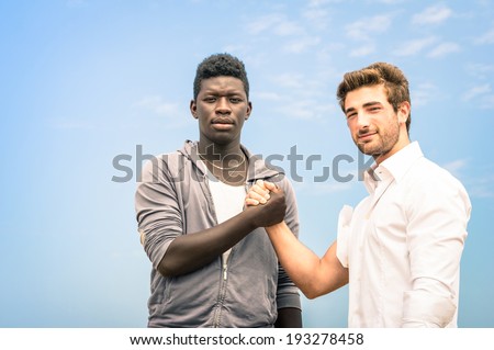 Afroamerican and caucasian men shaking hands in a modern handshake to show each other friendship and respect - Arm wrestling against racism on a blue sky