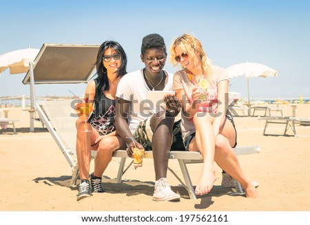 Happy multiracial girl friends drinking cocktails with tablet pc at the beach - Concept of modern vacations with girlfriends and afroamerican guy enjoying wifi internet in the summer