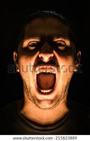 Horror scene with screaming scary human face with a harsh light on a black background - Halloween concept with young man with open mouth and teeth