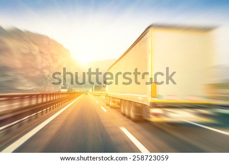 Generic semi trucks speeding on the highway at sunset - Transport industry concept with semitruck containers driving to the mountain pass - Warm editing with pop filtered sunshine and blurred edges
