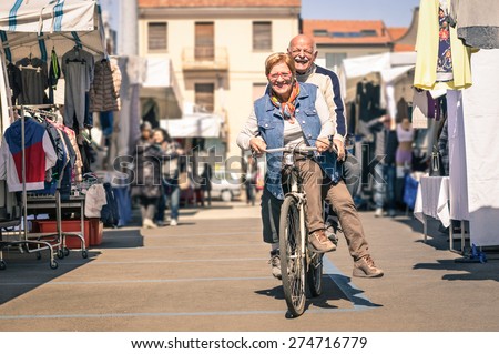 Happy senior couple having fun with bicycle at flea market - Concept of active playful elderly with bike during retirement - Everyday joy lifestyle without age limitation in a spring sunny afternoon
