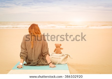 Young single woman sitting at beach with teddy bear looking at the sea - Solitude and loneliness concept with imaginary friendship and melancholic feelings - Warm vintage filter with enhanced sunshine
