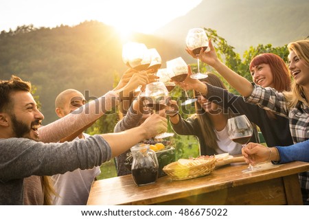 Happy friends having fun outdoors - Young people enjoying harvest time together at farmhouse vineyard countryside - Youth and friendship concept - Focus on hands toasting red wine glass with sun flare