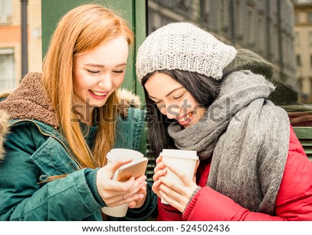 Happy girlfriends best friend having fun with coffee takeaway cup in autumn season - Friendship concept with joyful girls sharing time together with smartphone and winter clothes - Bright vivid filter