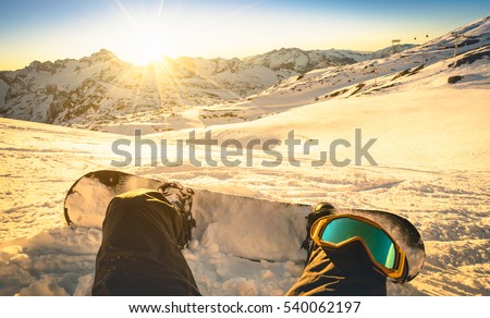 Snowboarder sitting on relax moment at sunset in Les Deux Alpes ski resort - Winter sport concept with person on top of the mountain ready to ride down - Legs view point with warm backlighting filter