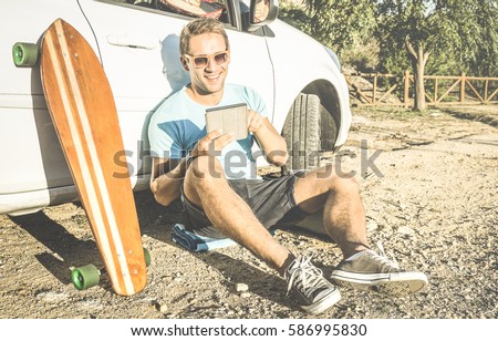 Young hipster fashion guy working remote on computer tablet sitting at car on road trip - New trend and technology concept with digital nomad lifestyle - Traveler man on retro contrasted filtered look