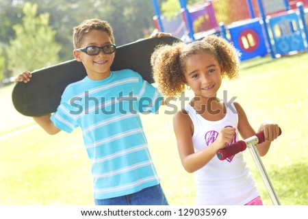 Full length portrait of cute little girl with push scooter and boy carrying skateboard over his shoulders at park. Horizontal shot.