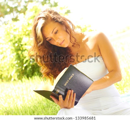 Pretty young woman reading bible in park. Horizontal shot.