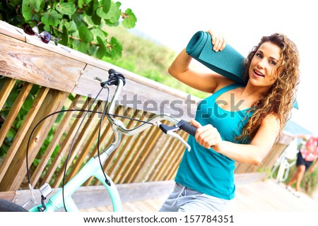 Bike riding female taking a break on boardwalk smiling holding yoga mat. Horizontal shot, copy space, color image, pretty yoga female on bike.