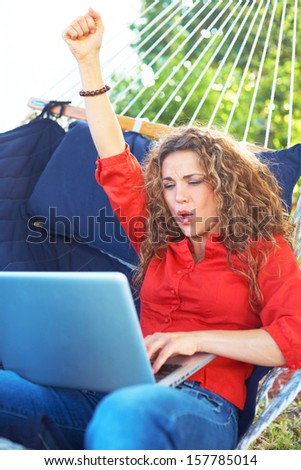 Excited beautiful female laying in hammock typing on laptop looking at screen.Vertical, color image, young woman on vacation relaxing in hammock with lap top,arm in air excited about some good news.