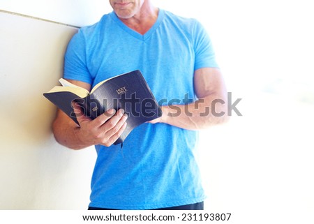 Midsection of mature man reading Bible while leaning on wall