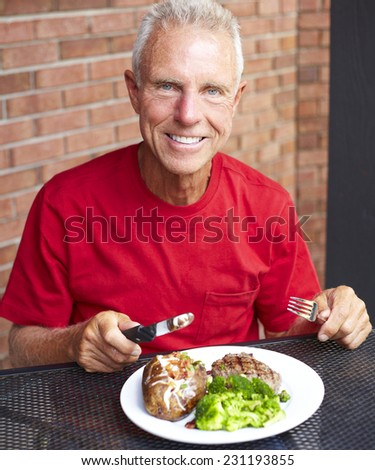 Portrait of happy senior man eating strip steak served with loaded baked potato and broccoli at restaurant table