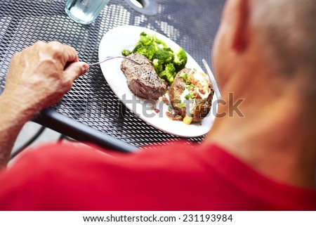 Cropped image of senior man eating strip steak served with loaded baked potato and broccoli at restaurant table