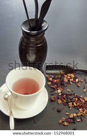 On a dark surface is white cup with fruit tea. Nearby stand and lie tea spoons and blades, also scattered pieces of fruit tea.