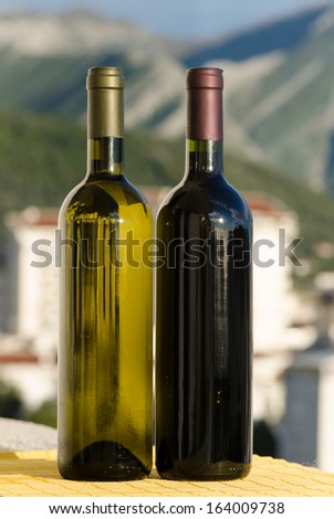A bottle of white wine and a bottle of red wine outside in sunny weather with a blurry mountain town on the background